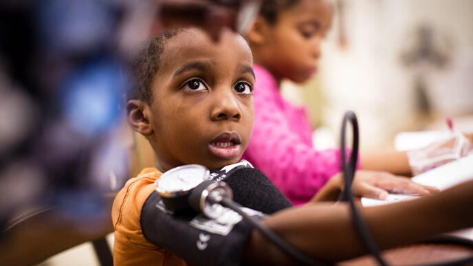 A young boy with a blood pressure cuff around his arm looks up at a health professional reading the dial.