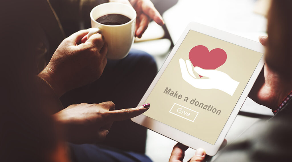 Several hands hold and point to a tablet device whose screen reads Make A Donation|Caleb Gardner and Kathleen Murphy stand at the front of a conference room