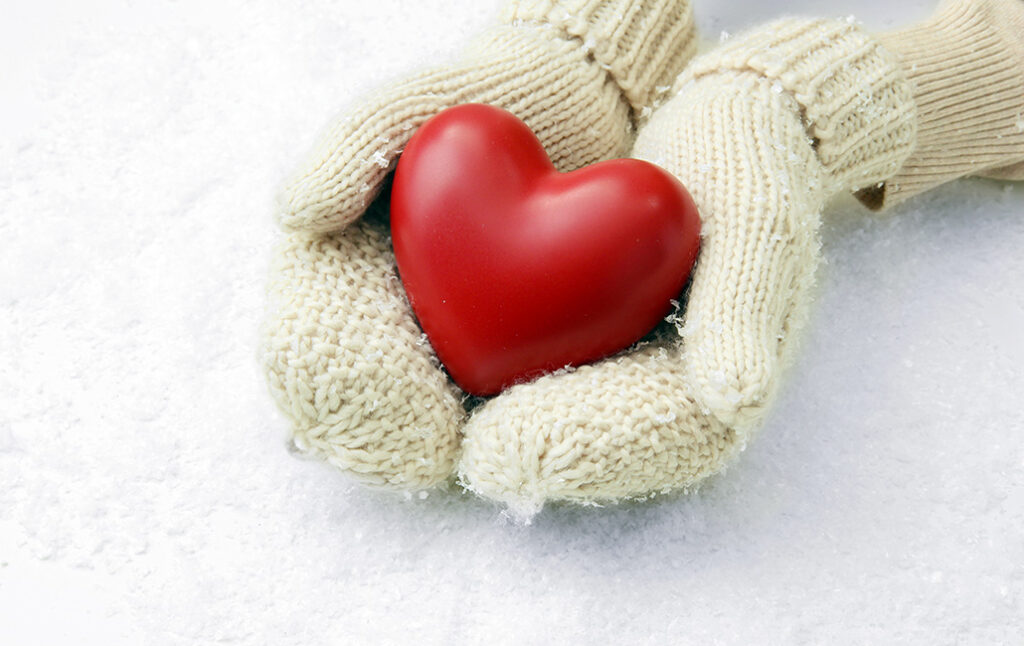 A pair of hands wearing mittens hold a red heart against a background of snow