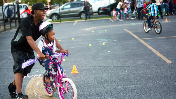 A young girl pedals a bike while a man holds the handlebars and helps her steer