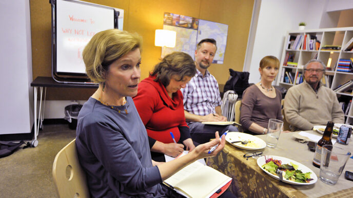 Group of people seated around a table talking|Whiteboard with the words Welcome to Why Not Local?|Closeup of metal picture frame and desk accessories|Susan Page Estes leaning forward listening to conversation|View from above of thirteen people seated around a table talking