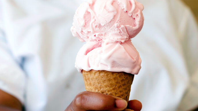 An ice cream server holding a cone with two scoops|Hand scooping ice cream from a freezer case|Jeni’s Storefront on Southport Avenue in Chicago|Three people sitting at a table holding waffle cones