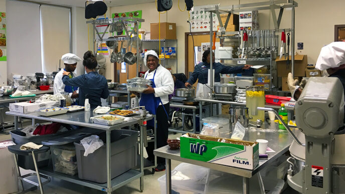A large institutional kitchen where five people wearing aprons and caps are working|Several students seen from behind in a greenhouse with potted plants in the foreground|Three young male students wearing safety glasses use hand tools in a wood shop|An instructor and a student work together to wash a white SUV