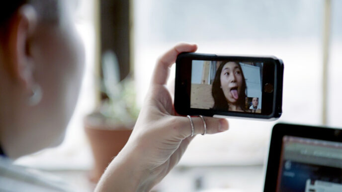 A young woman’s hand holding a phone to record herself on video; the screen shows her sticking her tongue out in a playful face