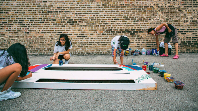 Three teenagers paint a large mural panel lying on the ground