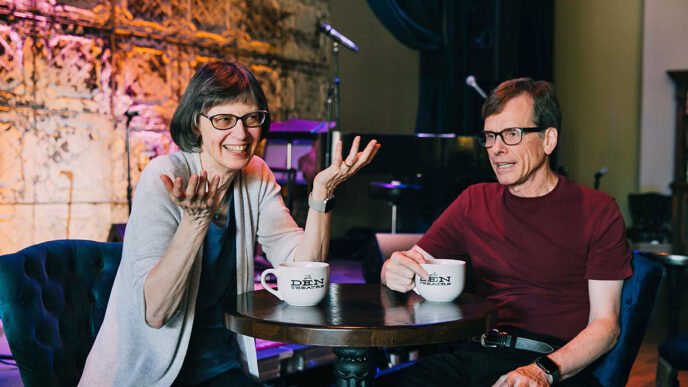 Two people sitting and talking at a table in front of a small stage.