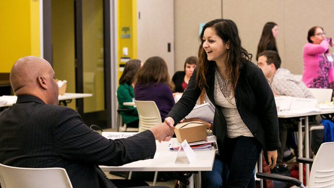 A standing woman smile sand shakes the hand of a man seated in a chair|A meeting room with people seated around shared tables.