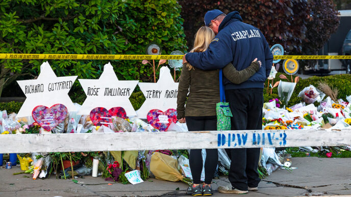 A man wearing a jacket that reads Chaplain embraces a woman as they stand in front of a row of memorials on a sidewalk|I Gave to #LoveIsStronger|The Pittsburgh Steelers team logo reworked to include a Star of David