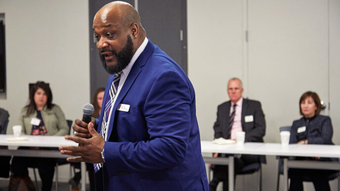 A man wearing a blue suit stands in front of a town of people seated at a conference room table.
