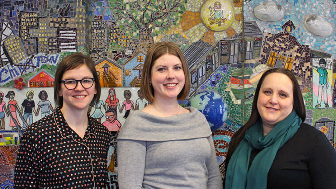 Three people stand in front of a brightly colored mosaic||Three people stand in front of a brightly colored mosaic.