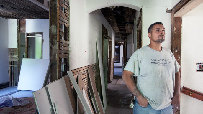 David Figueroa stands inside a vintage building under renovation|Figueroa supervises a crew of construction workers restoring a building foundation.