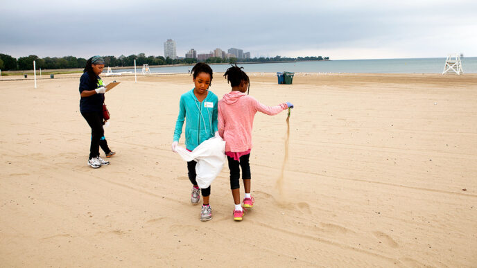 Two sisters pick up trash from the 63rd Street Beach during a cleanup event organized by the Honeycomb Project|A girl and her parents pick up trash at the 63rd Street Beach during a cleanup event organized by the Honeycomb Project|A family checks in to begin beach cleanup|Volunteers collect trash from the sand|A young child watches as girls test the water quality at a 63rd Street Beach cleanup event.
