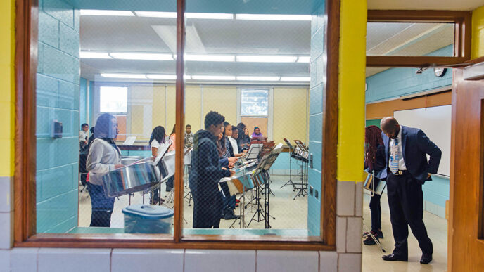 Students rehearse in a music classroom at Whistler Elementary School|A young musician holds a mallet poised over a steel drum|Julian Champion conducts a music class|Two girls in the percussion section read sheet music together.