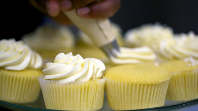 A baker pipes swirls of vanilla frosting onto a platter of cupcakes|Lisa Thompson serving up a cupcake and smiling|Lisa Thompson working alongside a trainee in the bakery's kitchen|.