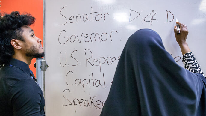 Mahjeed Marrow with a student from his citizenship class write on the classroom whiteboard|Mahjeed Marrow waits for a train on the Red Line station platform|A young boy plays a computer game in the office at the Pan-African Association|A student fills out a practice test of citizenship questions|Mahjeed Marrow helps a student complete a list of elected officials.