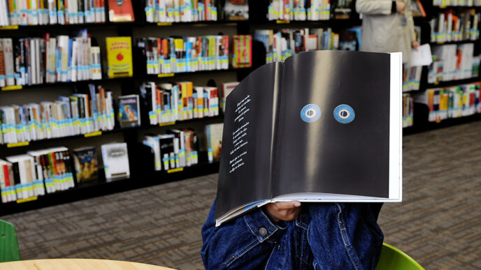 A boy sitting in a library holds a picture book in front of his face|Deborah Summers.