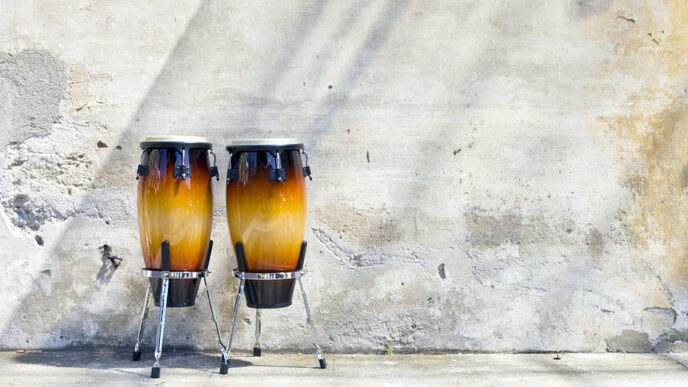 Two conga drums in front of an exterior wall|Carlos Cornier plays drums outside a storefront.