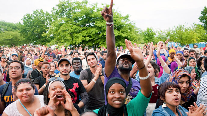 Rows of people seated in folding chairs and standing at an outdoor concert clap and cheer.