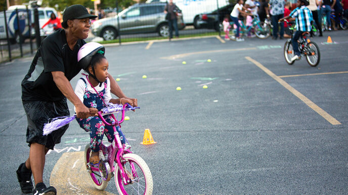 A young girl pedals a bike while a man holds the handlebars and helps her steer.