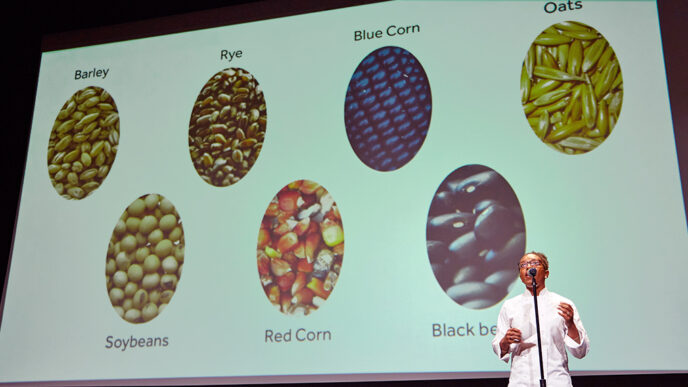 A chef gives a stage presentation about beans and grains|A display of garlic and root vegetables on a table|Four people on stage in front of a projection screen|Members of Team Leverage hugging and smiling|A group of judges seated at a table while one speaks into a microphone|Crowded auditorium at the Museum of Contemporary Art|.