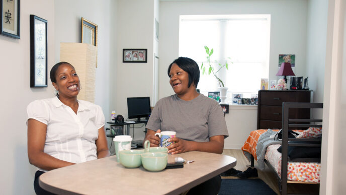 Two women drinking coffee at a kitchen table|A woman lying on a bed studying a textbook.