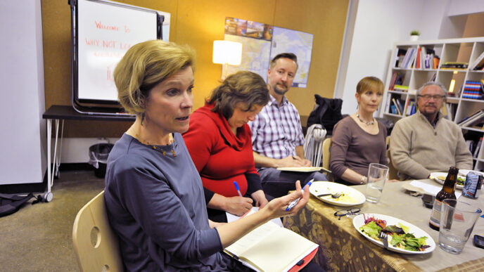 Group of people seated around a table talking|Whiteboard with the words Welcome to Why Not Local?|Closeup of metal picture frame and desk accessories|Susan Page Estes leaning forward listening to conversation|View from above of thirteen people seated around a table talking.