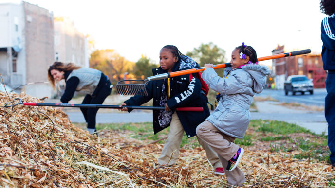 Two young girls raking wood chips in a garden|A colorfully painted stump with handprints and the words Black Lives Matter|A garden educator listens to a conversation|A young woman leans on a rake.