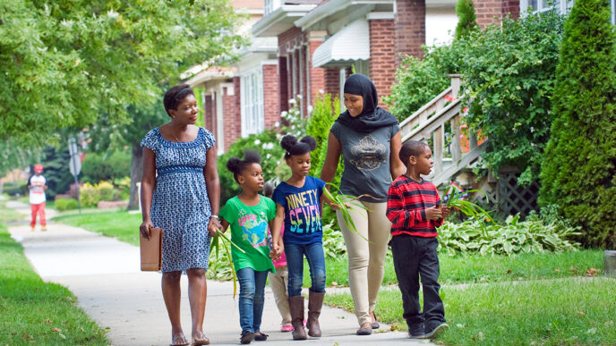 Two women and three young children walk along a sidewalk in a residential neighborhood|Two women seated at a table in conversation|A woman holds a toddler on her lap as they look out a window.