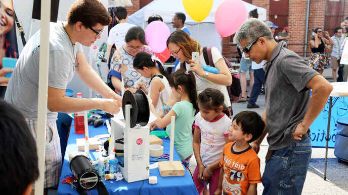 Families examine robotics components at an outdoor festival table.