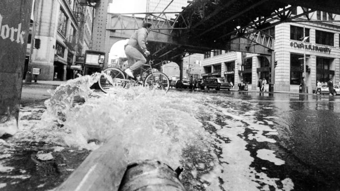 Cyclists pedal along a flooded street in the Loop as water gushes from pipes|.
