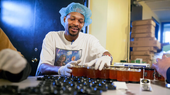 A man seated at a workbench seals jars of honey.