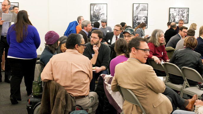 A group of people seated in chairs and motorized wheelchairs in an animated discussion|A speaker seated behind a table addresses a meeting using a microphone.