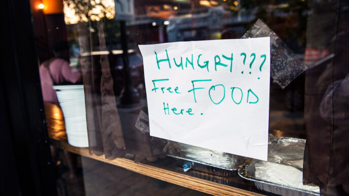 A hand-written sign taped on a restaurant window reads Hungry? Free Food Here|A A man seated at a table speaks and gestures to several women seated beside him||A woman tapes a sign onto the window of a restaurant|A smiling woman with her arms folded|Four people fill their plates from chafing dishes on a buffet table|A man gestures and speaks intensely while people in the background listen|A young woman speaks to a man seated at a table beside her.