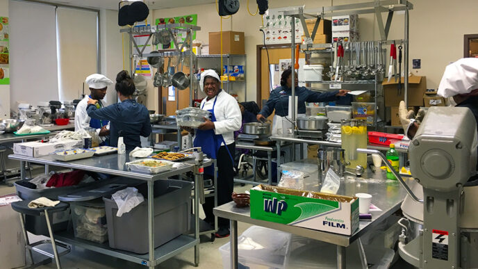 A large institutional kitchen where five people wearing aprons and caps are working|Several students seen from behind in a greenhouse with potted plants in the foreground|Three young male students wearing safety glasses use hand tools in a wood shop|An instructor and a student work together to wash a white SUV.