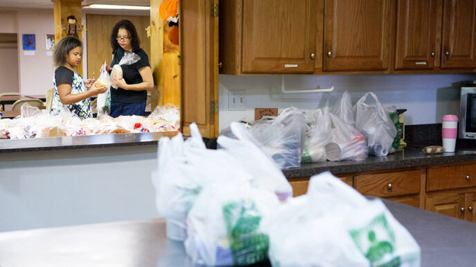 A woman and a young girl pick up packaged food in a large kitchen.