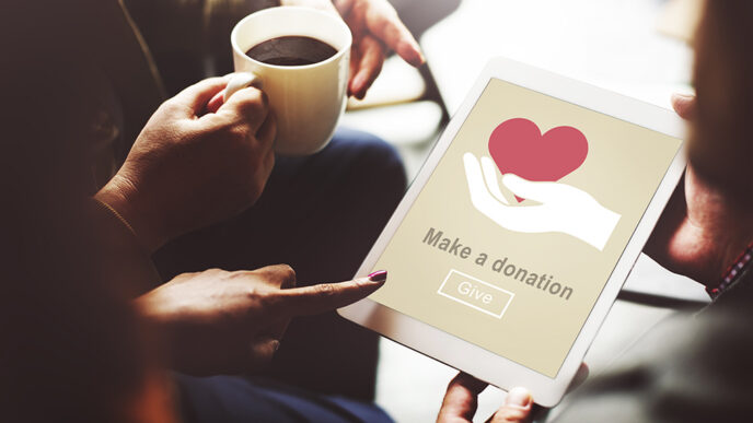 Several hands hold and point to a tablet device whose screen reads Make A Donation|Caleb Gardner and Kathleen Murphy stand at the front of a conference room.