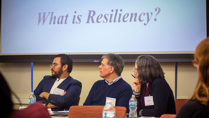 Three people steed below a projection screen displaying the words What Is Resiliency?|Several dozen people in a conference room face a row of panelists in conversation.