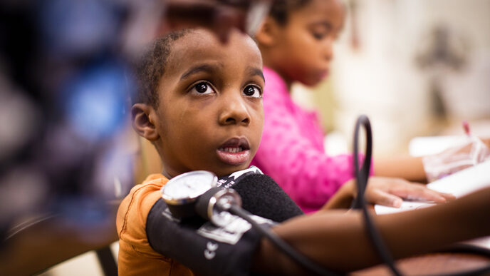 A young boy with a blood pressure cuff around his arm looks up at a health professional reading the dial.
