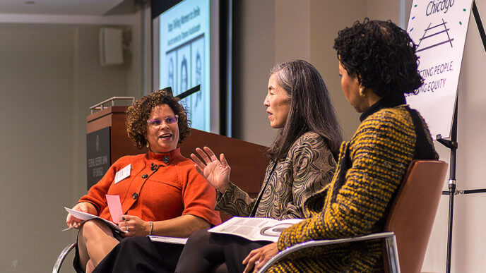 Three women sit in chairs in a stage.