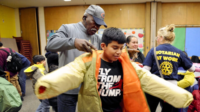 A man wearing a baseball cap helps a boy put on a new tan winter coat|A small girl sits at a table beside her mother.