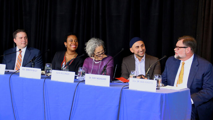 Five people seated behind a table for a panel discussion. The man seated at far right is speaking.