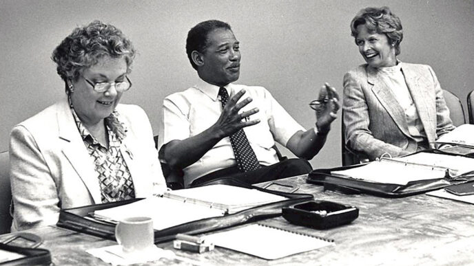 Black & white archival photo of a meeting at the Trust's offices. At a conference table covered with binders and papers.