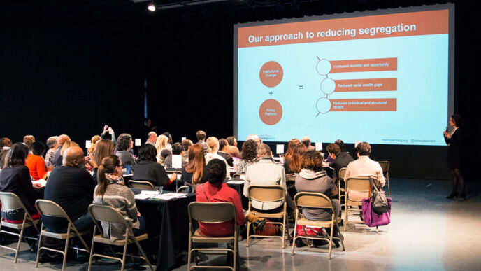 A room full of people in folding chairs face a presenter. A projection screen shows a slide that reads Our Approach to Reducing Segregation|A long table of people in conversation under bright stage lights|Five people sit at a table in conversation. Two are seated with their backs to us.