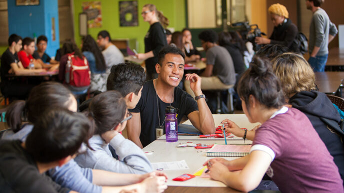 A smiling young man seated around a table with a group of high school students.