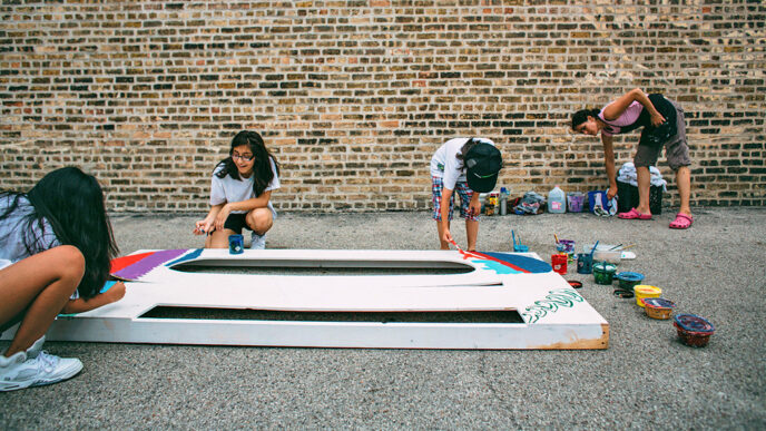 Three teenagers paint a large mural panel lying on the ground.