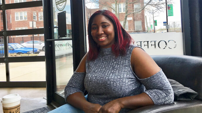 A woman seated on a leather sofa in a coffee shop smiles into the camera. Buildings are visible through the large windows behind her.