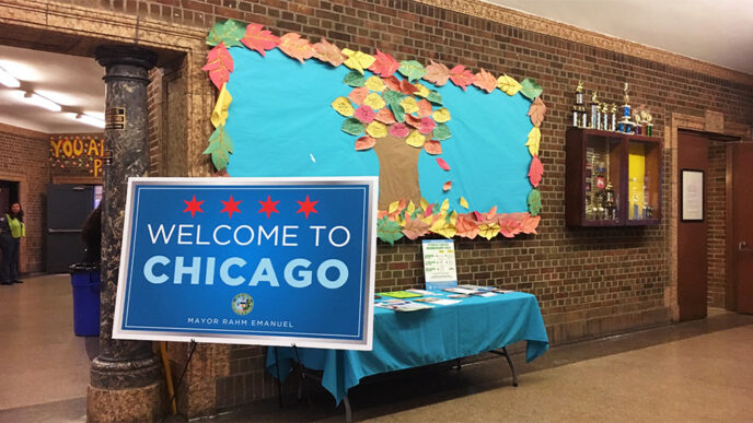 An easel in a large gymnasium holds a large sign reading Welcome to Chicago|A woman seated at a table speaks and gestures with her hands|Five people seated around a table in front of a projection screen.