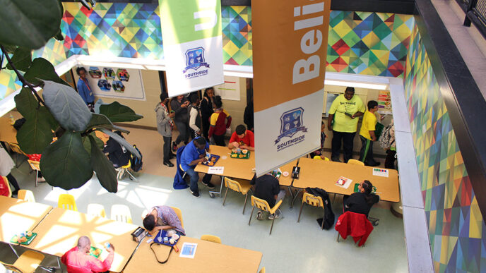 Aerial shot of the Southside Occupational Academy cafeteria. Students gather to have lunch and converse at the cafeteria tables.|A worker wearing a chef's jacket and sequined cap pushes a tray of ingredients through a commercial kitchen|Five people smile to the camera in front of a sign reading Southside Occupational Academy - Welcome.