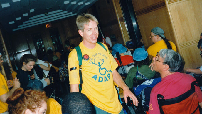 A man wearing a t-shirt that says ADAPT amid a group of people using wheelchairs|Vintage photo of protestors shouting and holding signs.