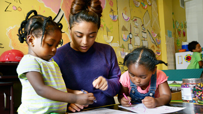 A young woman sits at a table between two small girls who are coloring with crayons